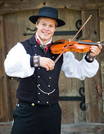En smilende ung mann i tradisjonell norsk bunad spiller fiolin foran en trevegg.