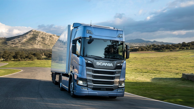 En blå Scania lastebil kjører på en vei med grønne åser i bakgrunnen.