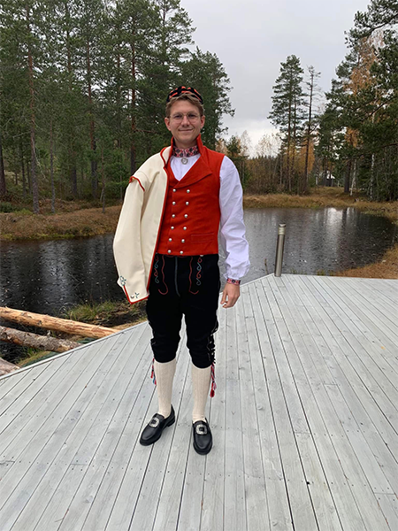 Mann i tradisjonell norsk bunad med rød vest og hvit skjorte, står på en brygge ved en innsjø om høsten.