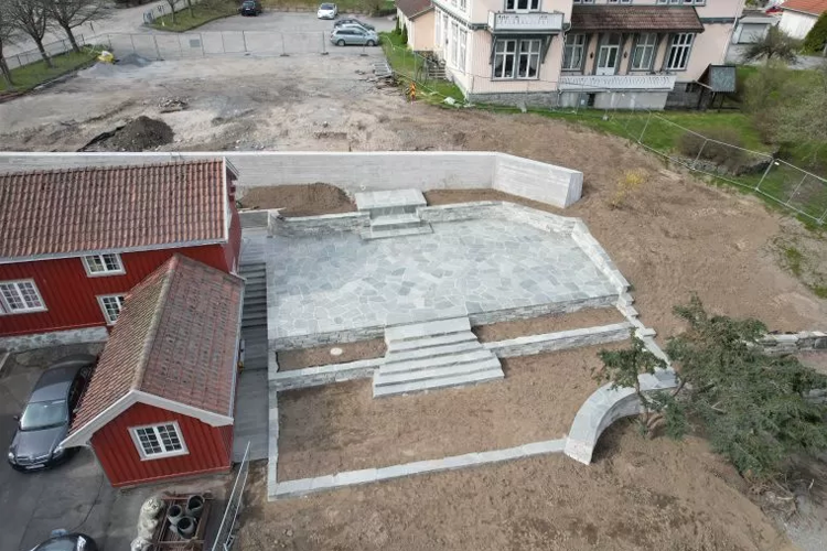 Utsikt over en nyanlagt steinlagt terrasse med trapper, omgitt av bygninger og tomt land.