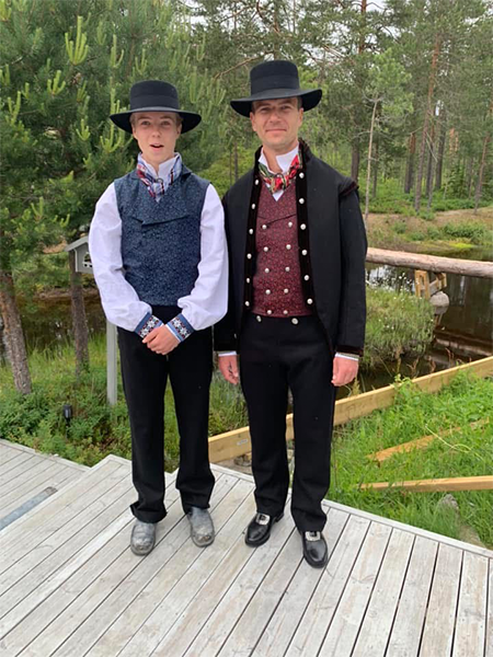 To unge menn iført tradisjonelle norske bunader med hatter, står på en tredekk ved en innsjø.
