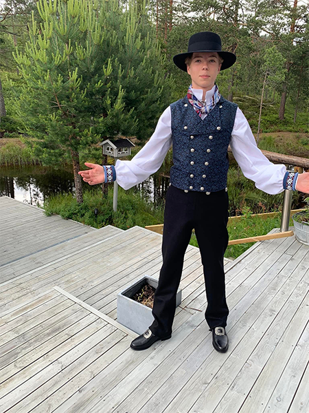 Ung mann i tradisjonell norsk bunad med vest og hatt, står på en tredekk med natur i bakgrunnen.