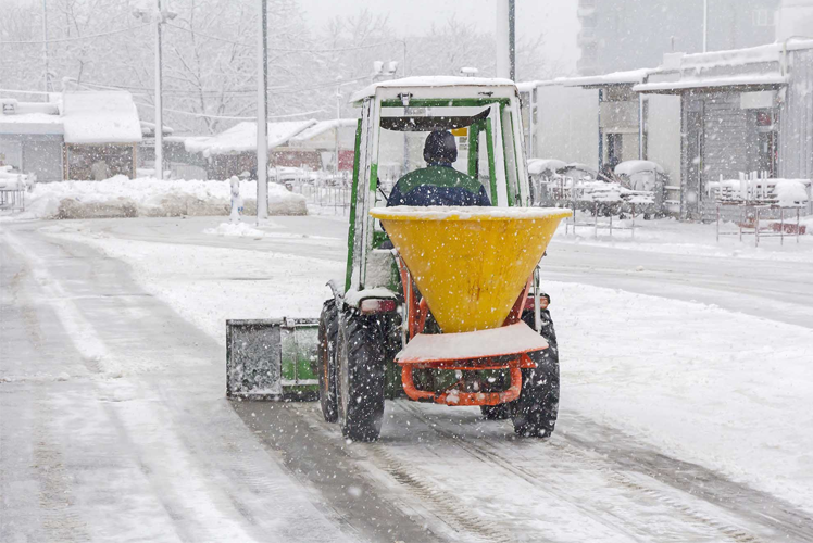 En traktor med snøplog og saltspreder kjører på en snødekt vei i snøvær.