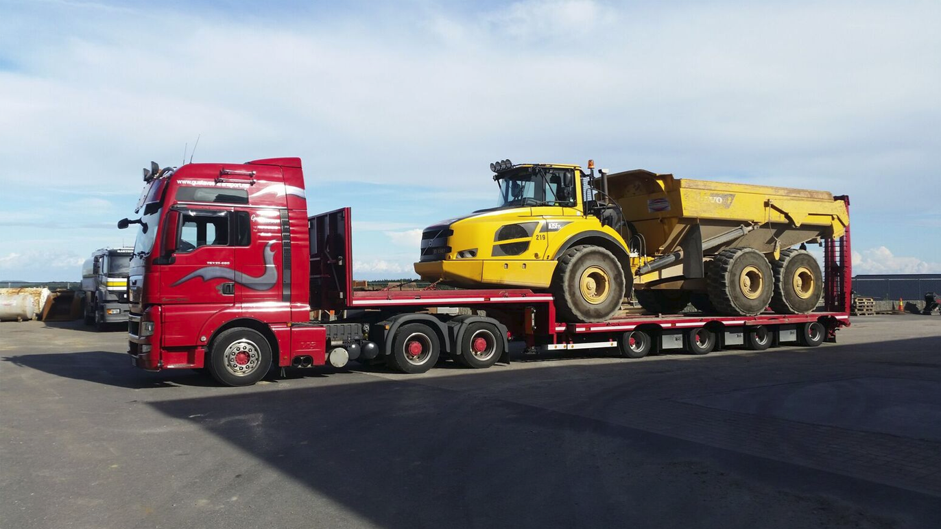En rød lastebil med en gul dumper på tilhenger, parkert på en byggeplass under en blå himmel.