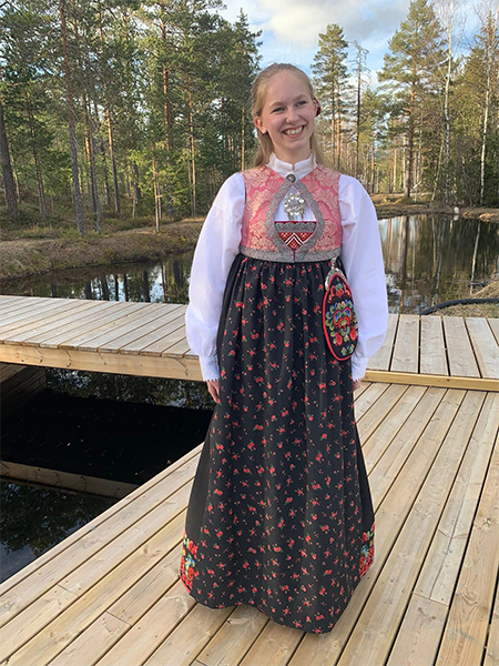 Ung kvinne i tradisjonell bunad med blomsterdesign, står på en treplattform ved en innsjø omringet av trær.