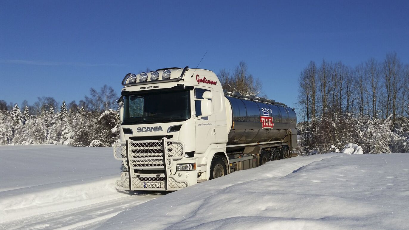 En hvit Scania tankbil kjører gjennom snødekt landskap med trær i bakgrunnen.