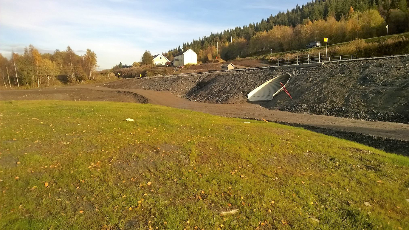 Landskap med grønt gress, grusvei og en hvit avløpstunnel, omgitt av trær og hus i bakgrunnen.