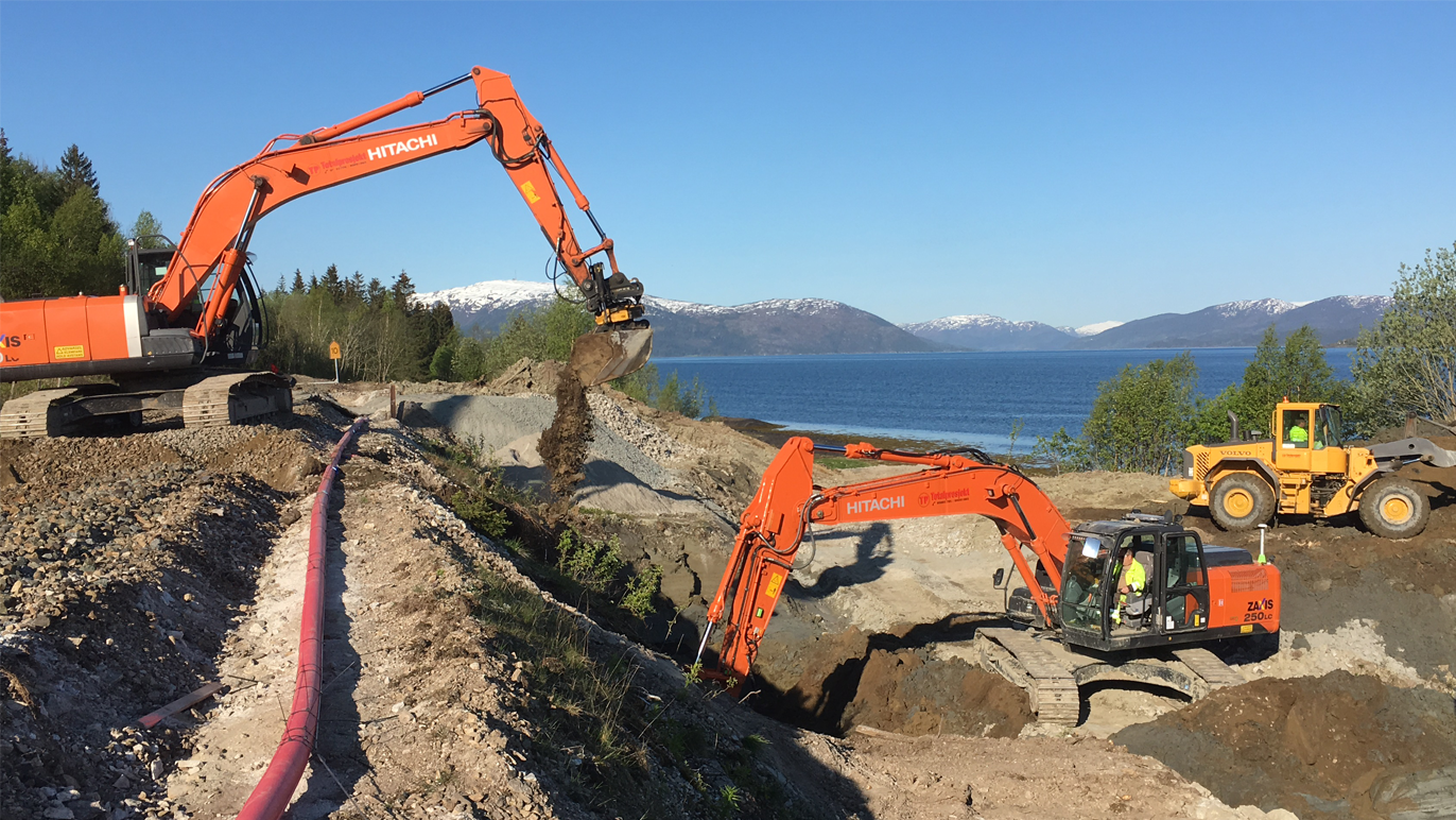 To store og en gravemaskin arbeider med graving ved en vei nær en innsjø, med fjell i bakgrunnen.