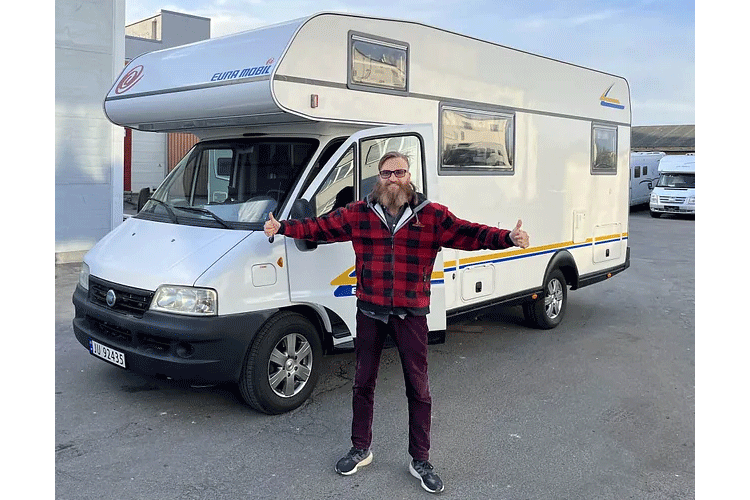 En mann med skjegg står foran en campingvogn og gir tommel opp, iført rutete skjorte og jeans.