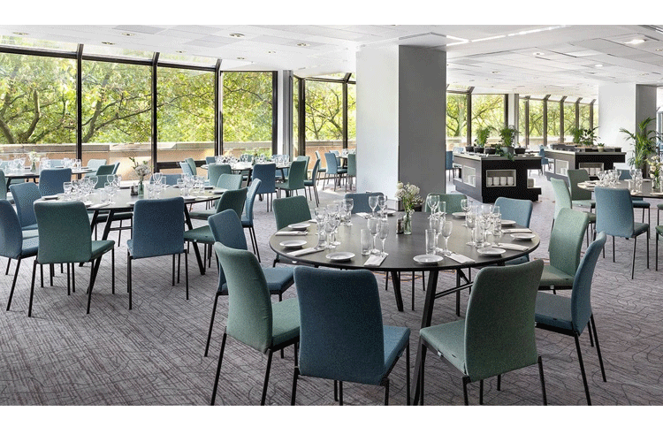 Moderne restaurant med runde bord, blå stoler og store vinduer som gir naturlig lys.