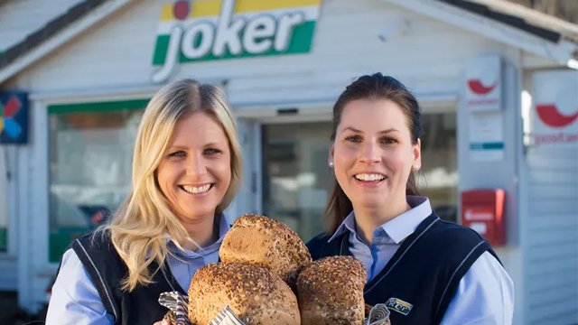 To kvinner smiler og holder en haug med brød foran Joker-butikken.
