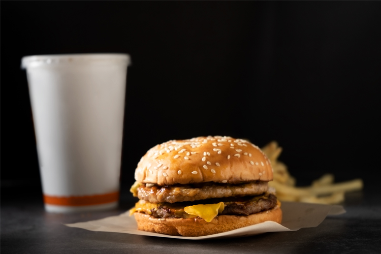 En saftig hamburger med sesamfrøbrød, ost og to kjøttpattyer, med en brus og pommes frites i bakgrunnen.