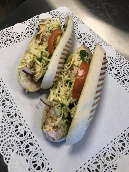 To brød med pølse, salat, tomat, løk og ost, servert på en hvit serviett med dekorativ kant.