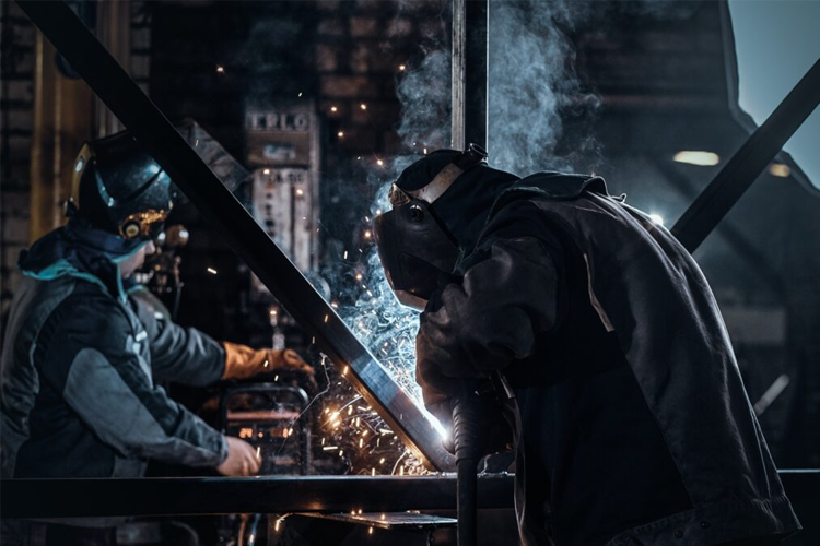 To personer som sveiser i en industriell setting, med gnister og damp som fyller luften.