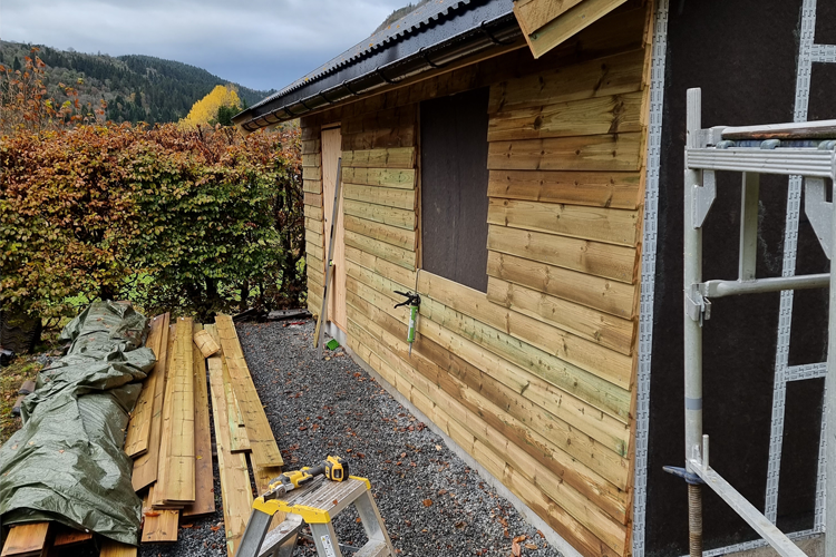 Ny trevegg under bygging med uferdige planker og verktøy på bakken, omringet av høstfarger.
