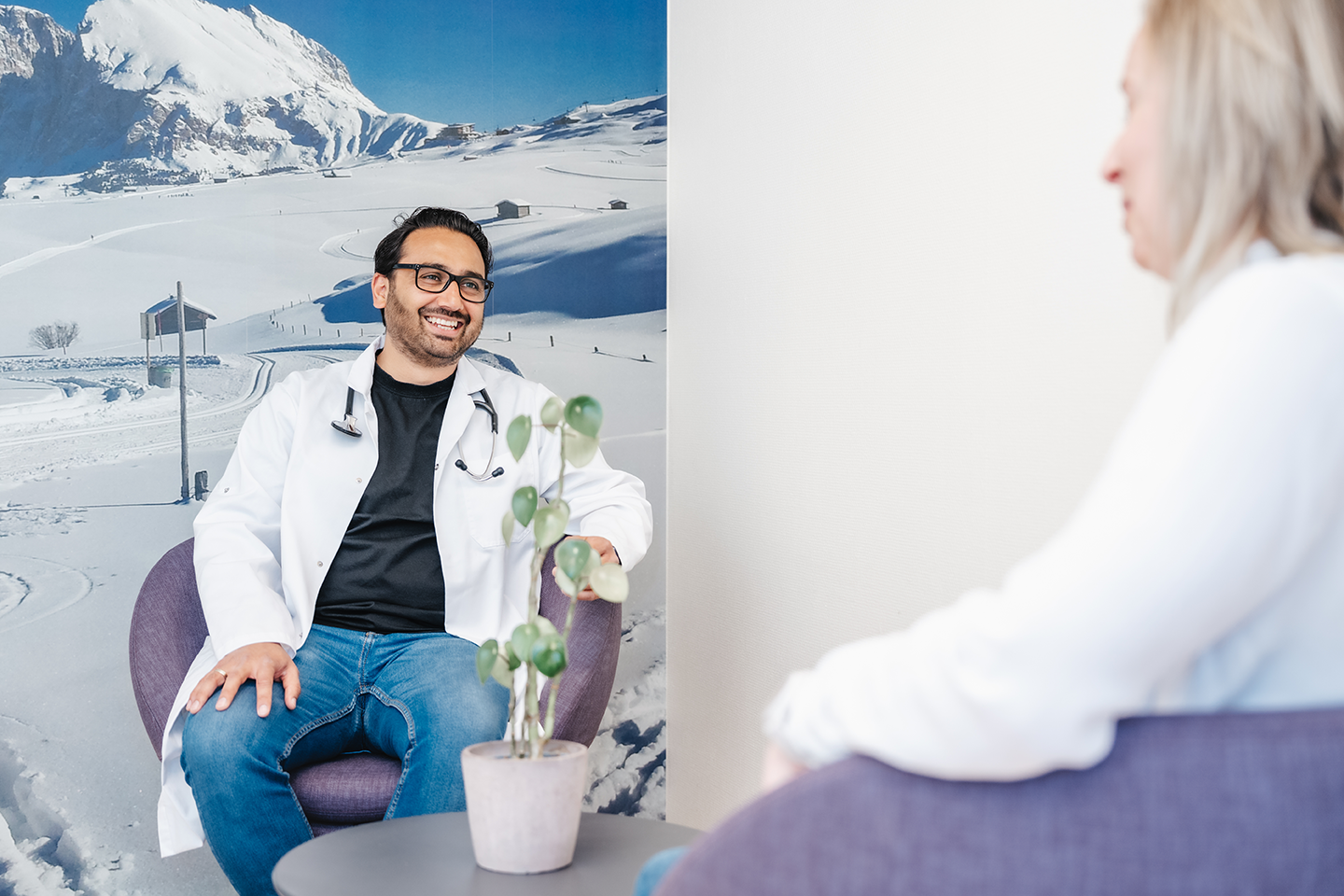 Lege i hvit frakk smiler mens han snakker med en pasient i en moderne konsultasjonsstol.