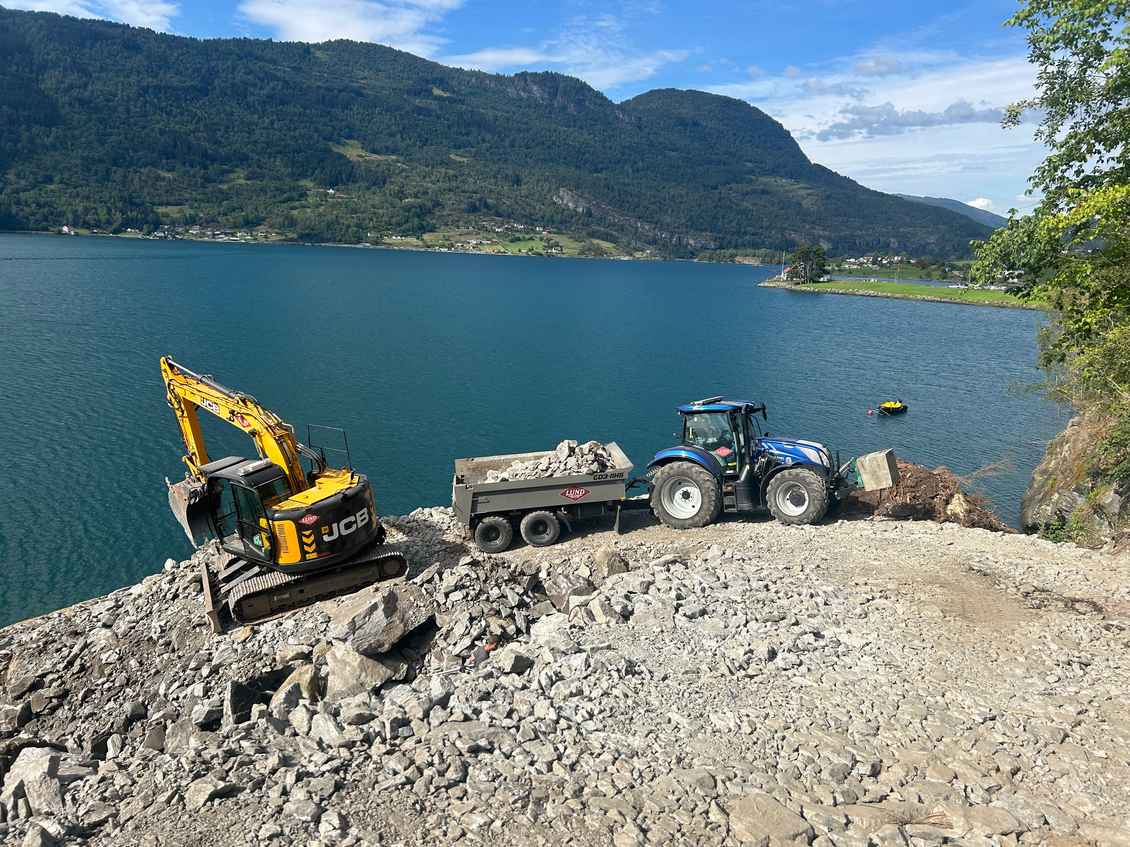 Gravemaskin og traktor med tilhenger arbeider på en steinete strand ved en innsjø med fjell i bakgrunnen.