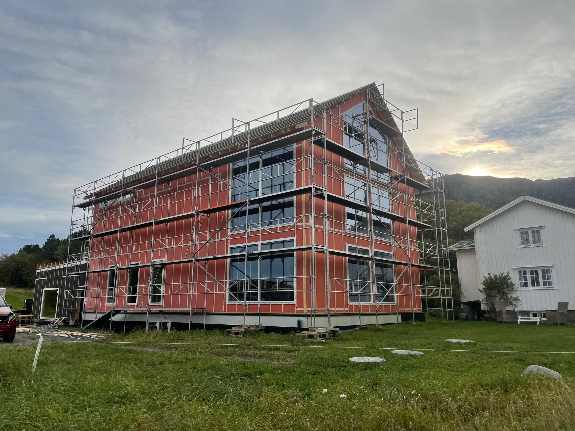 Nybygget hus med stillas, store vinduer og moderne design, omgitt av grønt gress og fjell i bakgrunnen.