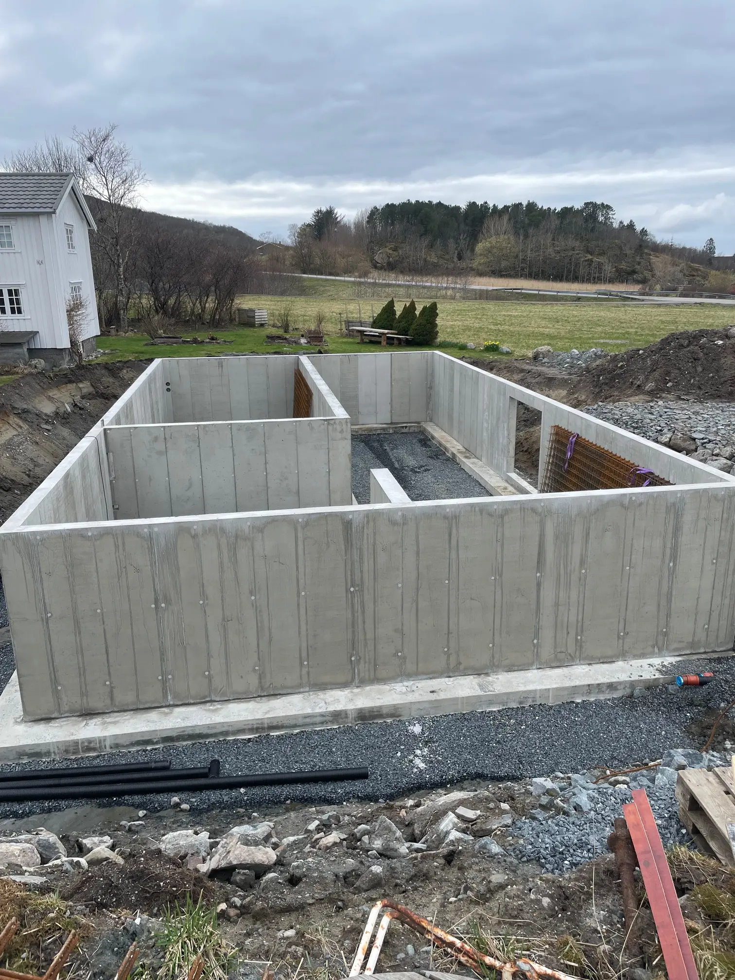 Fundamentet til et hus med betongvegger, omringet av grus og landskap med trær i bakgrunnen.