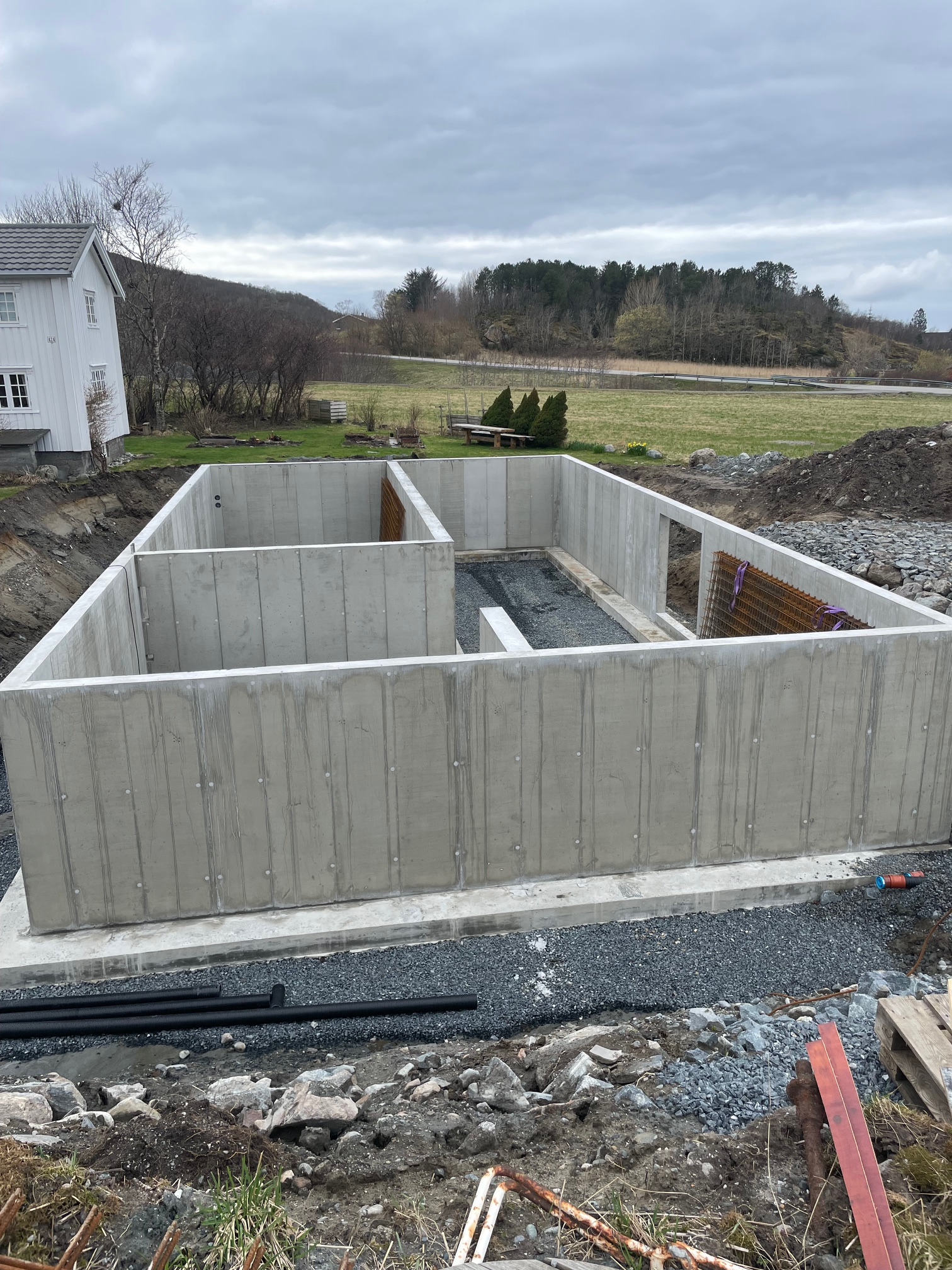 Fundamentet til et hus med betongvegger, omringet av grus og landskap med trær i bakgrunnen.