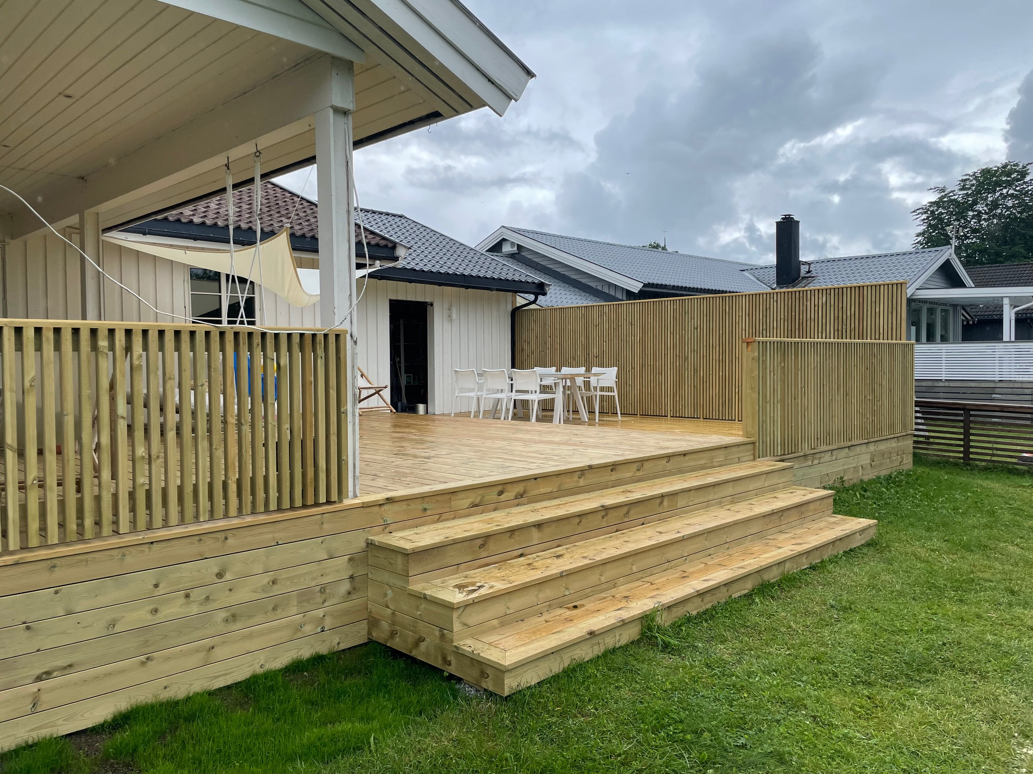Terrasse med tretrapper, stoler og bord, omgitt av gjerde, med hus i bakgrunnen under en skyet himmel.