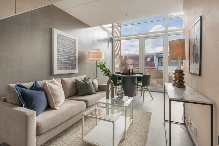 Moderne stue med sofa, bord og store vinduer som slipper inn naturlig lys. Dekorative puter og planter gir liv til rommet.