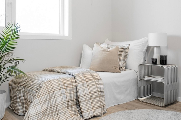 Lyst soverom med en enkel seng, rutete sengetøy, puter, lampe og en liten hylle med planter.