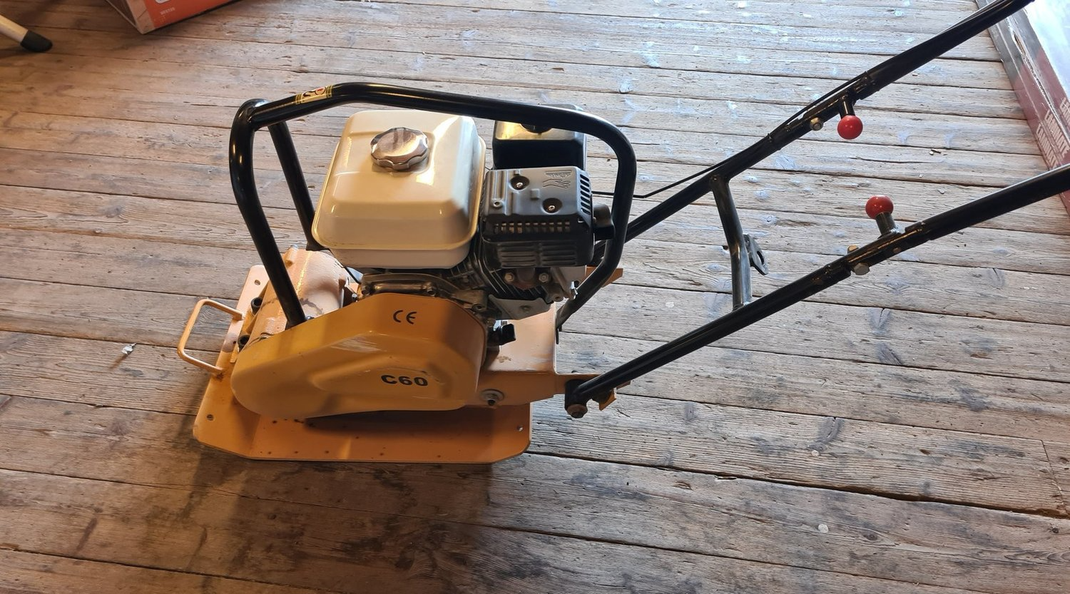En gul vibroplate med motor, plassert på et tregulv i et verksted.