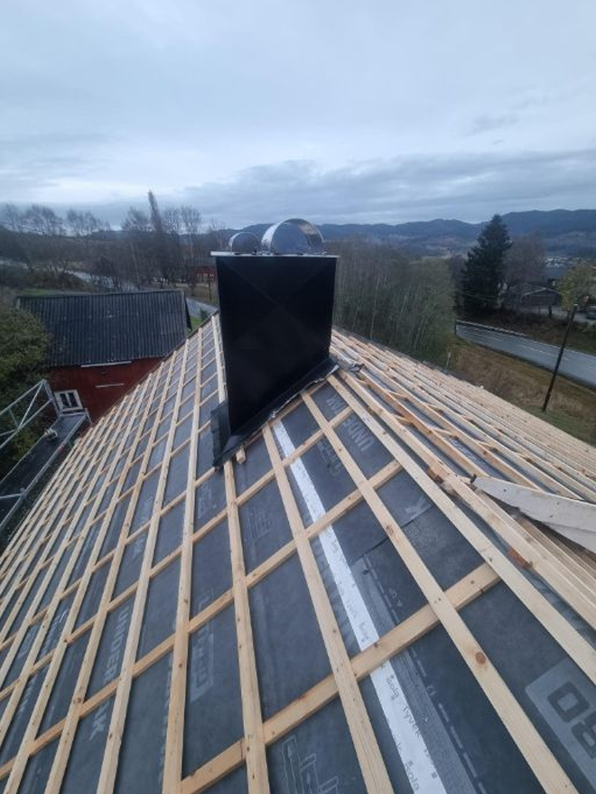 Tak med takstein og ventilasjonsrør, sett fra oven, med grå himmel og landskap i bakgrunnen.