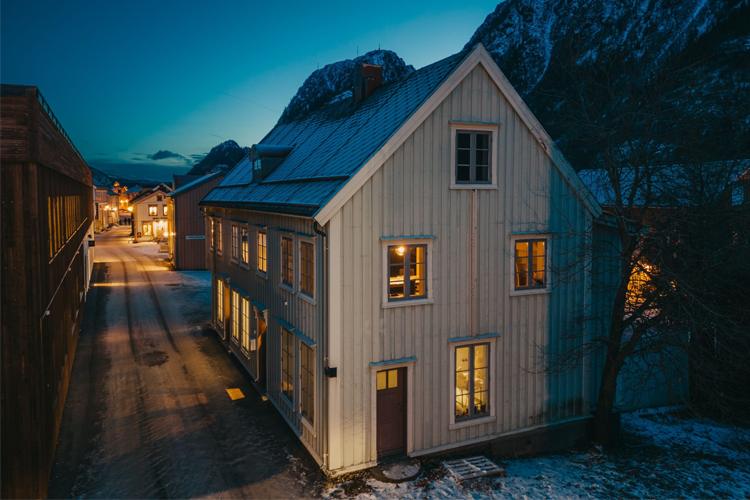 Et hvitt trehus med mange vinduer, beliggende i en snødekt gate ved foten av fjellene om kvelden.
