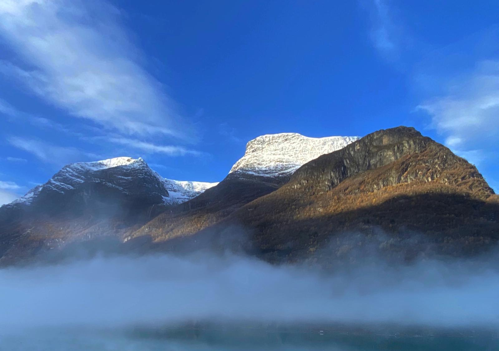 Fjell med snødekte topper og tåke som dekker dalen under en klar blå himmel.