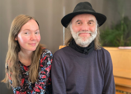 To personer som smiler mot kameraet, en kvinne med langt hår og en fargerik topp, og en mann med langt grått hår og hatt.
