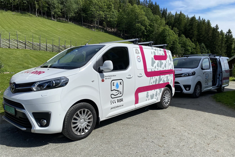 To hvite varebiler med logoer parkert på en grusvei, omringet av grønn natur og trær.
