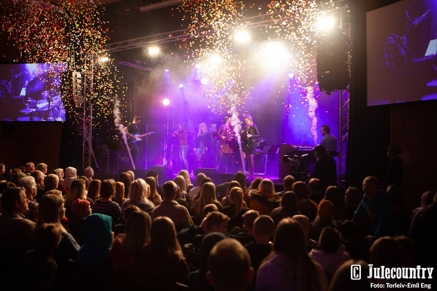 En konsert med band på scenen, publikum sitter i mørket, konfetti faller fra taket under lysene.