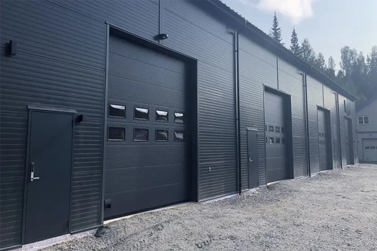 En moderne garasje med flere store porter og en liten dør, omgitt av grus og trær i bakgrunnen.
