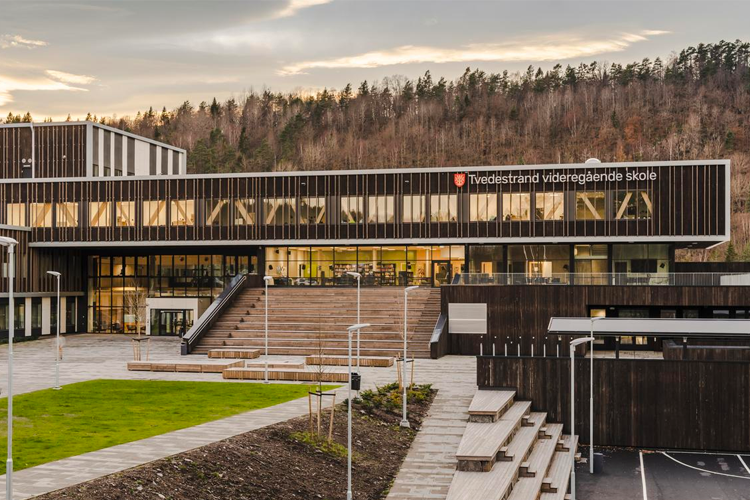Moderne skolebygning med trepanel og store vinduer, omgitt av grøntområde og trappetrinn.