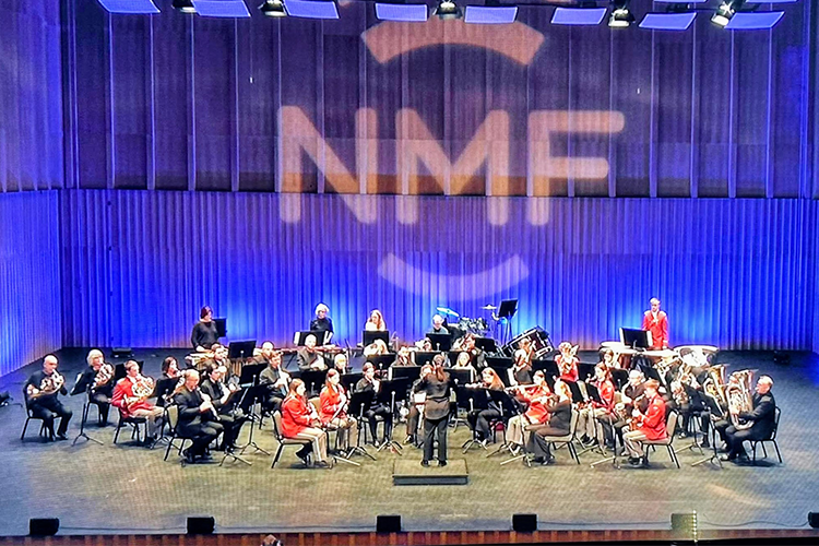 En stor musikkorpsopptreden på scenen med musikere i røde skjorter og dirigent foran.