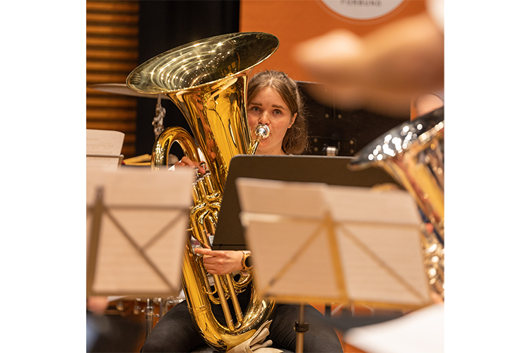 En ung kvinne spiller tuba i et orkester, omgitt av noteark og musikere.