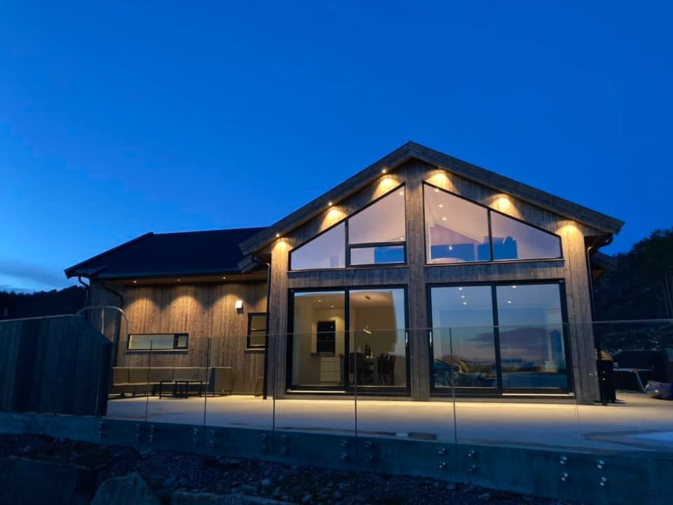 Moderne hytte med store vinduer og terrasse, beliggende ved kysten under en blå himmel.
