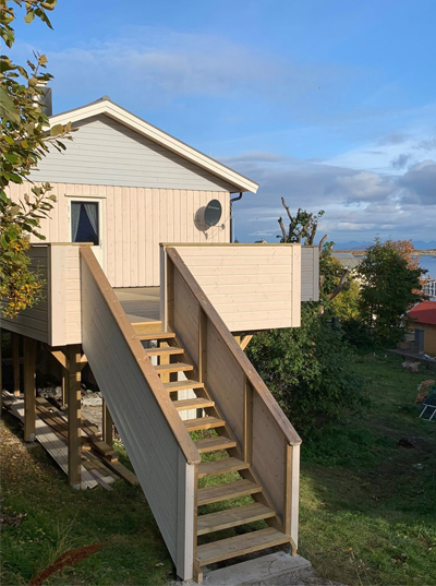 Hus med tretrapp og veranda, plassert på en høyde med utsikt over landskapet og himmelen.