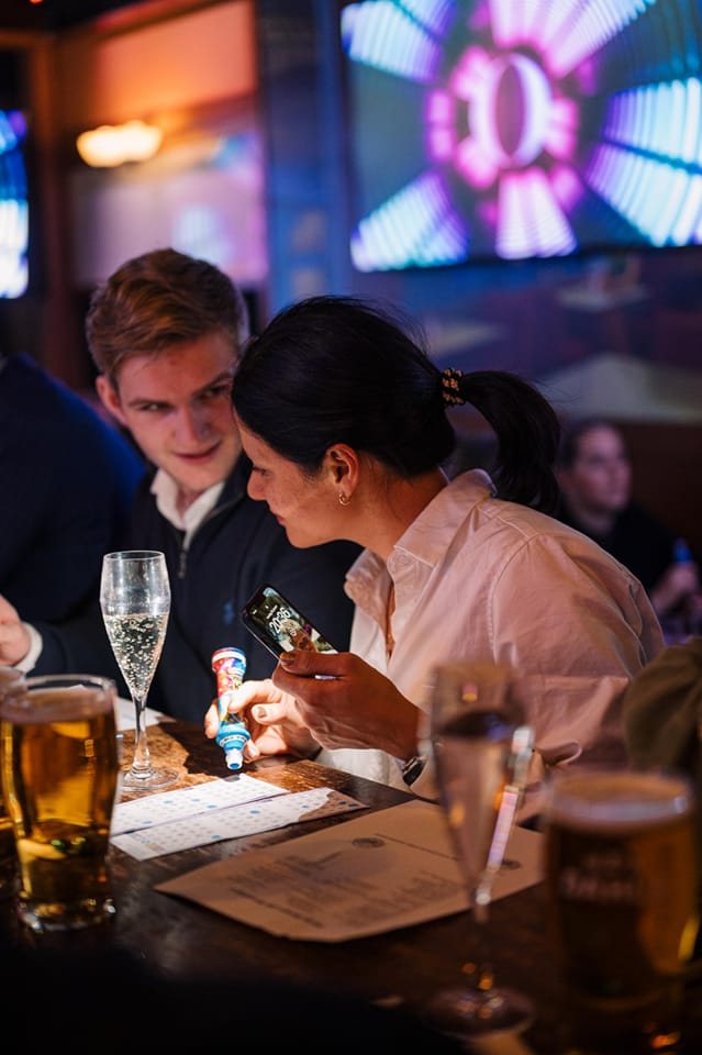 To personer sitter ved et bord på en bar, med drinker og en lekefigur mellom dem. Skjermer i bakgrunnen lyser opp.