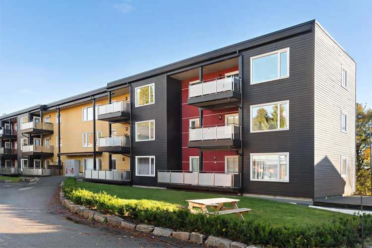 Moderne leilighetsbygg med balkonger, omgitt av grøntområde og grusvei. Solfylt dag med klar himmel.
