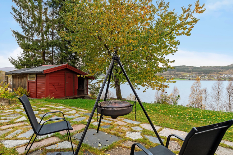 Utsikt over en hage med en grill, to stoler, en rød hytte og en innsjø i bakgrunnen.