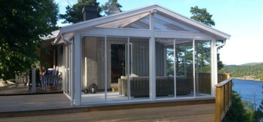 Moderne hytte med glassveranda og utsikt over vannet, omgitt av trær og en treterrasse.