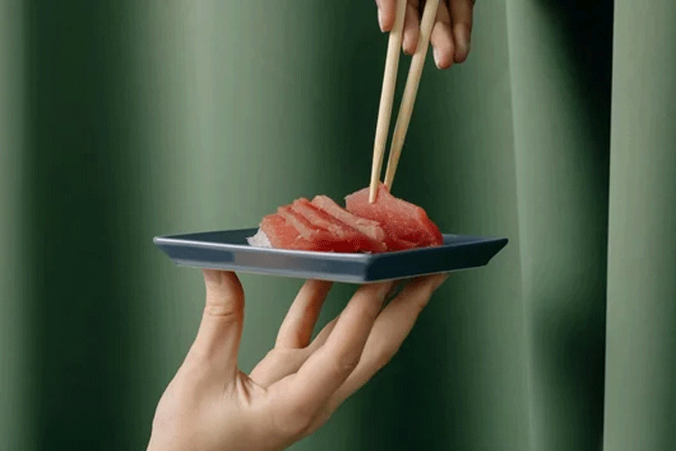 Hånd som holder en tallerken med sushi, mens en annen hånd bruker spisepinner for å ta en bit.