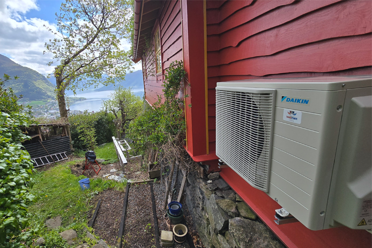 Utendørs varmepumpe montert på en rød husvegg med utsikt over fjorden og trær i bakgrunnen.