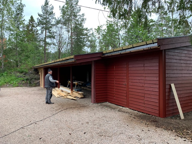En mann står foran en rød garasje med trepanel, omgitt av trær og gruslagt vei.