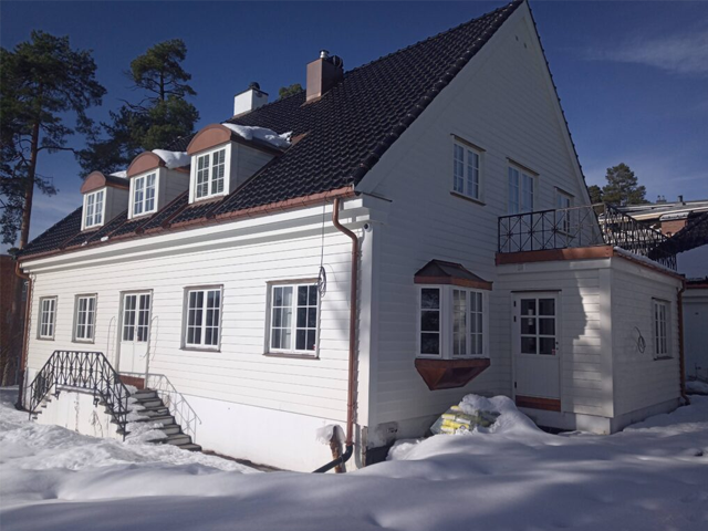 Et stort, hvitt hus med skråtak og flere vinduer, omgitt av snø og trær.