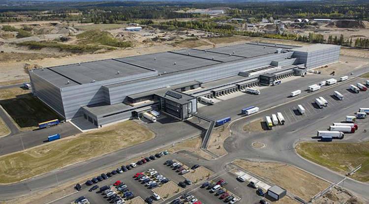 Moderne lagerbygning med lastebiler og parkerte biler på området, omgitt av et industrielt landskap.
