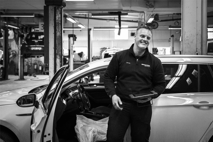 En smilende mekaniker står ved siden av en bil i et verksted, med verktøy i hånden og en arbeidsplass i bakgrunnen.